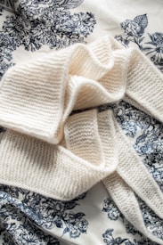 A soft, knitted cream-colored scarf arranged elegantly on a patterned fabric background. The fabric features intricate floral designs in black or dark blue, creating a striking contrast with the scarf.