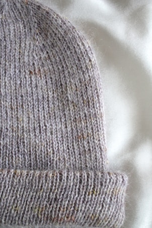 A close-up view of a knitted woolen hat lying on a soft, light-colored fabric. The hat has a ribbed texture with a folded brim, showcasing a speckled pattern with hints of brown and other muted colors.