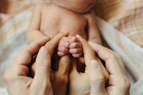 Hands gently cradling a newborn baby's tiny hands, conveying a sense of love and protection. The scene is intimate and tender.