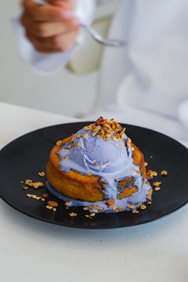 A scoop of purple ice cream is served on top of a warm, golden-brown pastry. The ice cream is melting slightly, creating a colorful drizzle over the edges of the dessert. It is garnished with granola and nuts, adding texture and contrast to the dish. A person in a white attire is holding a spoon in the background.