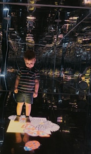 A young child stands in a room with reflective walls and ceiling, creating a kaleidoscopic effect. The child is dressed in a striped shirt and shorts, and looks down at a brightly lit pattern on the floor that resembles flowers and abstract shapes.