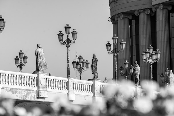 A black and white photograph features a bridge adorned with multiple statues standing on pedestals. Ornate street lamps are spaced between the statues, adding a historic or classical aesthetic. In the background, a large building with grand columns suggests monumental architecture.