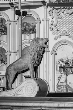 A stone sculpture of a lion stands atop a decorative pedestal, with its mouth open as if roaring. The background features ornate architecture with elaborate designs, columns, and arches. There's also an image of a playing card king and queen visible on a poster behind the sculpture.