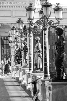 A series of classical statues line a bridge or walkway, flanked by ornate street lamps. The architecture features a traditional style with columns and decorative crowns on the lamps. Shadows from the lamps and statues create patterns on the ground. A building in the background has architectural details and inscriptions.