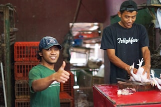 Two men are in a market or butcher shop. One man is wearing a green shirt and a cap, smiling and giving a thumbs up. The other man is focused on preparing a chicken on a chopping block, wearing a black shirt.