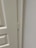 A close-up view of a door hinge on a light-colored door and frame. The door has a textured surface with panel detailing.