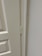 A close-up view of a door hinge on a light-colored door and frame. The door has a textured surface with panel detailing.