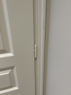 A close-up view of a door hinge on a light-colored door and frame. The door has a textured surface with panel detailing.