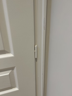 A close-up view of a door hinge on a light-colored door and frame. The door has a textured surface with panel detailing.