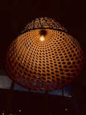 A large woven wicker lampshade hangs from the ceiling, illuminated by a single light bulb at its center. The intricate pattern of the wicker creates a geometric design with light and shadow effects, setting a warm and rustic ambiance.