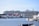 A large cruise ship is docked near a waterside town. The ship is massive, with multiple decks and identifiable signage on its side. Various smaller buildings with red and white exteriors are visible along the water's edge. Smaller boats are anchored nearby, adding to the maritime atmosphere.