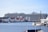 A large cruise ship is docked near a waterside town. The ship is massive, with multiple decks and identifiable signage on its side. Various smaller buildings with red and white exteriors are visible along the water's edge. Smaller boats are anchored nearby, adding to the maritime atmosphere.