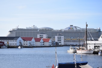 A large cruise ship is docked near a waterside town. The ship is massive, with multiple decks and identifiable signage on its side. Various smaller buildings with red and white exteriors are visible along the water's edge. Smaller boats are anchored nearby, adding to the maritime atmosphere.