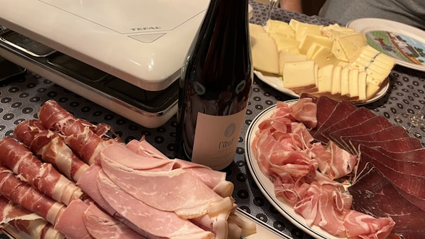 A table is set with a variety of charcuterie and cheese. There are neatly arranged slices of prosciutto, ham, and other cured meats, next to a bottle of wine. Adjacent is a plate of sliced cheese, ranging from mild to aged. The setup is completed with an electric grill in the background, suggesting preparation for a meal.