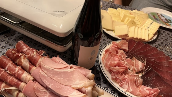 A table is set with a variety of charcuterie and cheese. There are neatly arranged slices of prosciutto, ham, and other cured meats, next to a bottle of wine. Adjacent is a plate of sliced cheese, ranging from mild to aged. The setup is completed with an electric grill in the background, suggesting preparation for a meal.