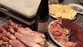 A table is set with a variety of charcuterie and cheese. There are neatly arranged slices of prosciutto, ham, and other cured meats, next to a bottle of wine. Adjacent is a plate of sliced cheese, ranging from mild to aged. The setup is completed with an electric grill in the background, suggesting preparation for a meal.