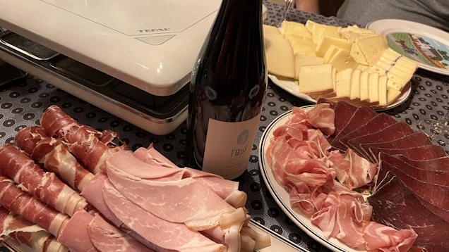 A table is set with a variety of charcuterie and cheese. There are neatly arranged slices of prosciutto, ham, and other cured meats, next to a bottle of wine. Adjacent is a plate of sliced cheese, ranging from mild to aged. The setup is completed with an electric grill in the background, suggesting preparation for a meal.