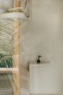 A minimalist interior featuring a smooth white wall, a light-colored cushioned seating area, and a small dark vase with a green plant placed on a white ledge. There is a glass reflection showing some palm leaves, adding a natural element to the scene.