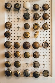 A collection of vintage doorknobs is mounted on a pegboard for display. The doorknobs vary in style and color, featuring intricate designs and patinas, with some appearing polished and others more worn. The arrangement is orderly in rows and columns.