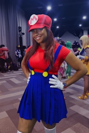 A person is dressed in a colorful costume, featuring a red shirt, matching red cap with a white emblem, and a blue skirt with suspenders. They are standing confidently with hands on hips, surrounded by others in costumes in a well-lit, indoor setting. The background includes people in colorful attire and a curtain, indicating a lively atmosphere.