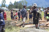 A group of people are gathered in an outdoor setting with grassy terrain and fallen logs. They are wearing a mix of casual clothing and traditional attire, and some are carrying backpacks. In the background, there are small houses and lush green trees with high mountains visible in the distance.