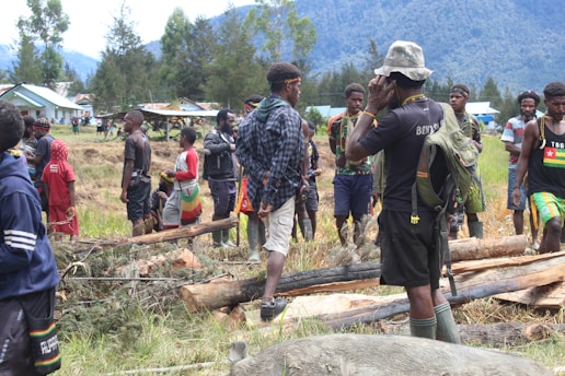 A group of people are gathered in an outdoor setting with grassy terrain and fallen logs. They are wearing a mix of casual clothing and traditional attire, and some are carrying backpacks. In the background, there are small houses and lush green trees with high mountains visible in the distance.