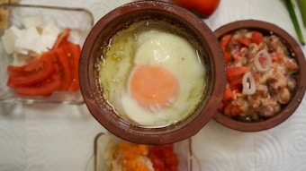 A cooked egg is placed in a rustic clay bowl, surrounded by various dishes including sliced tomatoes, cheese, and a mixture featuring beans and onions. The arrangement is on a paper towel, suggesting a casual, home-cooked meal.