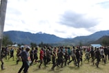 A large group of people are running together across a grassy area, surrounded by a scenic landscape of mountains and dense clouds. The participants are dressed in a variety of colorful clothing, suggesting a communal or cultural event, possibly a race or celebration.