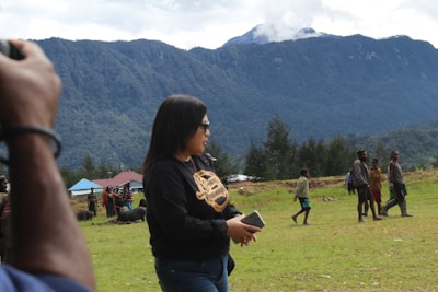 A group of people is gathered in an open grassy area with mountains in the background, some are walking while others are interacting in small groups. In the foreground, a person is holding a phone. Trees are scattered throughout the scene, and there are a few buildings with colorful roofs visible in the distance.