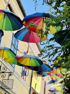Colorful umbrellas hang above a street, creating a vibrant canopy. The umbrellas are in various bright colors and contrast against the light-colored buildings and the blue sky. Leaves from nearby trees add a touch of greenery.
