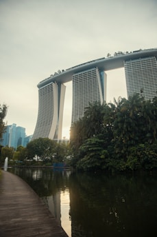 A futuristic-looking hotel with three tall towers connected by a large roof terrace stands prominently in the background. In the foreground, there is a serene body of water bordered by lush greenery and a clean, curved walkway. The scene is tranquil with a few modern buildings visible in the distance.