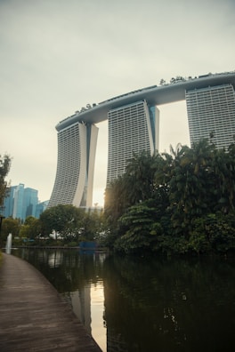 A futuristic-looking hotel with three tall towers connected by a large roof terrace stands prominently in the background. In the foreground, there is a serene body of water bordered by lush greenery and a clean, curved walkway. The scene is tranquil with a few modern buildings visible in the distance.