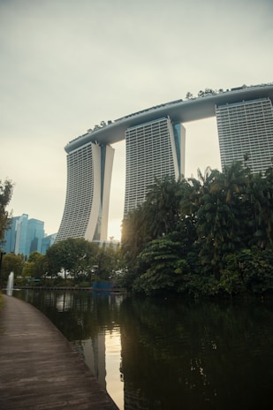 A futuristic-looking hotel with three tall towers connected by a large roof terrace stands prominently in the background. In the foreground, there is a serene body of water bordered by lush greenery and a clean, curved walkway. The scene is tranquil with a few modern buildings visible in the distance.