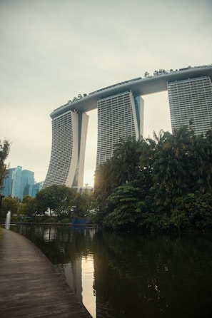 A futuristic-looking hotel with three tall towers connected by a large roof terrace stands prominently in the background. In the foreground, there is a serene body of water bordered by lush greenery and a clean, curved walkway. The scene is tranquil with a few modern buildings visible in the distance.