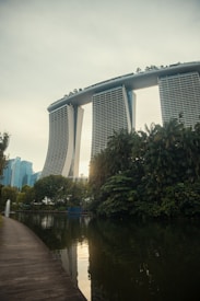 A futuristic-looking hotel with three tall towers connected by a large roof terrace stands prominently in the background. In the foreground, there is a serene body of water bordered by lush greenery and a clean, curved walkway. The scene is tranquil with a few modern buildings visible in the distance.