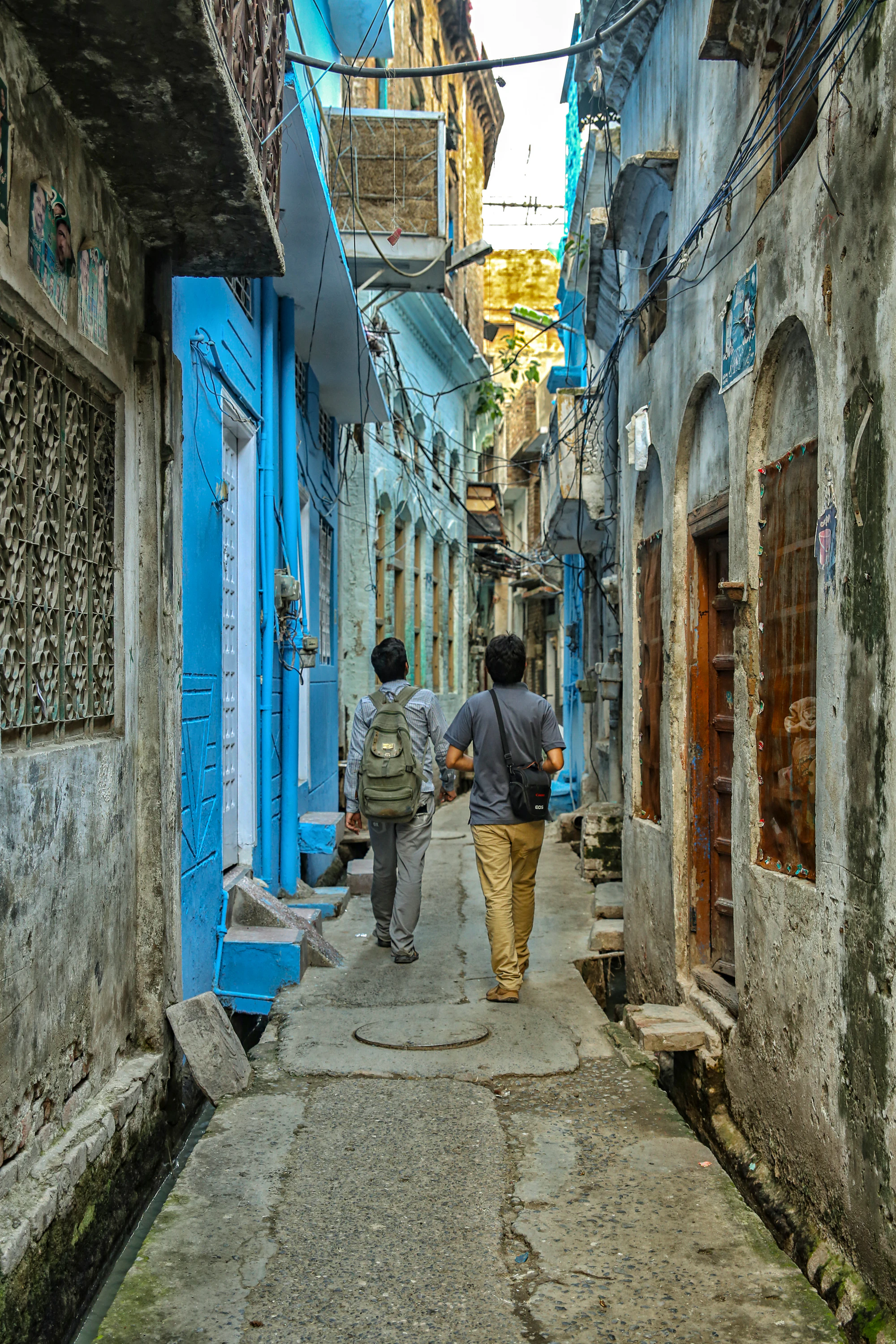 Two people walking away down a narrow sunlit alley between old brick houses in Pakistan