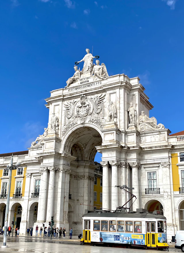 The Rua Augusta Arch with a yellow tram passing beneath it against a bright blue sky