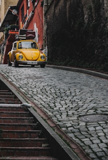 A vintage yellow Volkswagen Beetle is parked on a steep cobblestone street. It has luggage strapped to the roof and is positioned near a red building with a sign that reads 'Vintage Cafe.' The street is flanked by worn stone walls, and there is a set of stairs adjacent to the road.