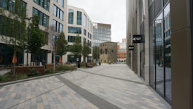 A modern urban setting featuring tall office buildings with large glass windows and clean, geometric lines. The walkway is lined with trees, plants, and wooden benches, creating a blend of nature and architecture. The scene is contemporary, with people walking and an older stone structure positioned centrally, contrasting with the modern buildings.