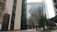 Modern urban area with tall glass buildings surrounding an open courtyard. Trees with autumn foliage are scattered throughout the courtyard, where a few people are seated on benches. The buildings feature a mix of architectural styles, with large glass facades and concrete structures.