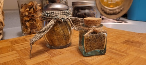 A wooden surface is adorned with two small glass jars filled with spices. One jar has a metal lid with a gingham fabric tied around it, and the other has a cork stopper with twine. In the background, other jars and kitchen items are partially visible.