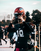 A football player wearing a maroon helmet and black uniform with the number 19 printed on the chest is adjusting his helmet on the field. Other players and people can be seen in the blurry background, indicating that they are on a football field with trees in the distance.