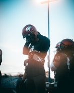 A group of football players, wearing helmets and uniforms, stands together in a field. One player is adjusting their helmet under the bright sky, with the sun creating a lens flare effect. The scene captures the pre-game preparation and focus of the athletes.