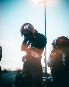 A group of football players, wearing helmets and uniforms, stands together in a field. One player is adjusting their helmet under the bright sky, with the sun creating a lens flare effect. The scene captures the pre-game preparation and focus of the athletes.
