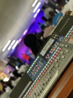 A digital audio mixing console with various colorful buttons and sliders is in the foreground, while a group of people is blurred in the background. The setting appears to be an indoor event with dynamic lighting.