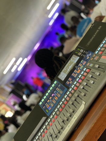 A digital audio mixing console with various colorful buttons and sliders is in the foreground, while a group of people is blurred in the background. The setting appears to be an indoor event with dynamic lighting.
