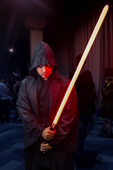 A person in a dark hooded robe holds a glowing red lightsaber in a dimly lit setting. The individual appears to be participating in a cosplay event, surrounded by other people in costumes.
