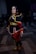 A person is dressed in detailed, vibrant cosplay attire, inspired by a character from a fantasy or animated series. The costume is elaborate, featuring a dark tunic with yellow and pink trim, a belt, and boots. The setting appears to be an indoor cosplay or comic convention with people in the background.