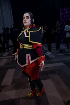 A person is dressed in detailed, vibrant cosplay attire, inspired by a character from a fantasy or animated series. The costume is elaborate, featuring a dark tunic with yellow and pink trim, a belt, and boots. The setting appears to be an indoor cosplay or comic convention with people in the background.