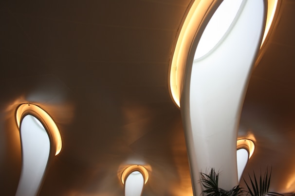 A modern architectural interior features curved, elongated light structures embedded in the ceiling. The lights are surrounded by a warm glow that contrasts with the smooth, dark surface of the ceiling. A few green plant leaves are visible at the bottom, adding a touch of nature to the sleek design.